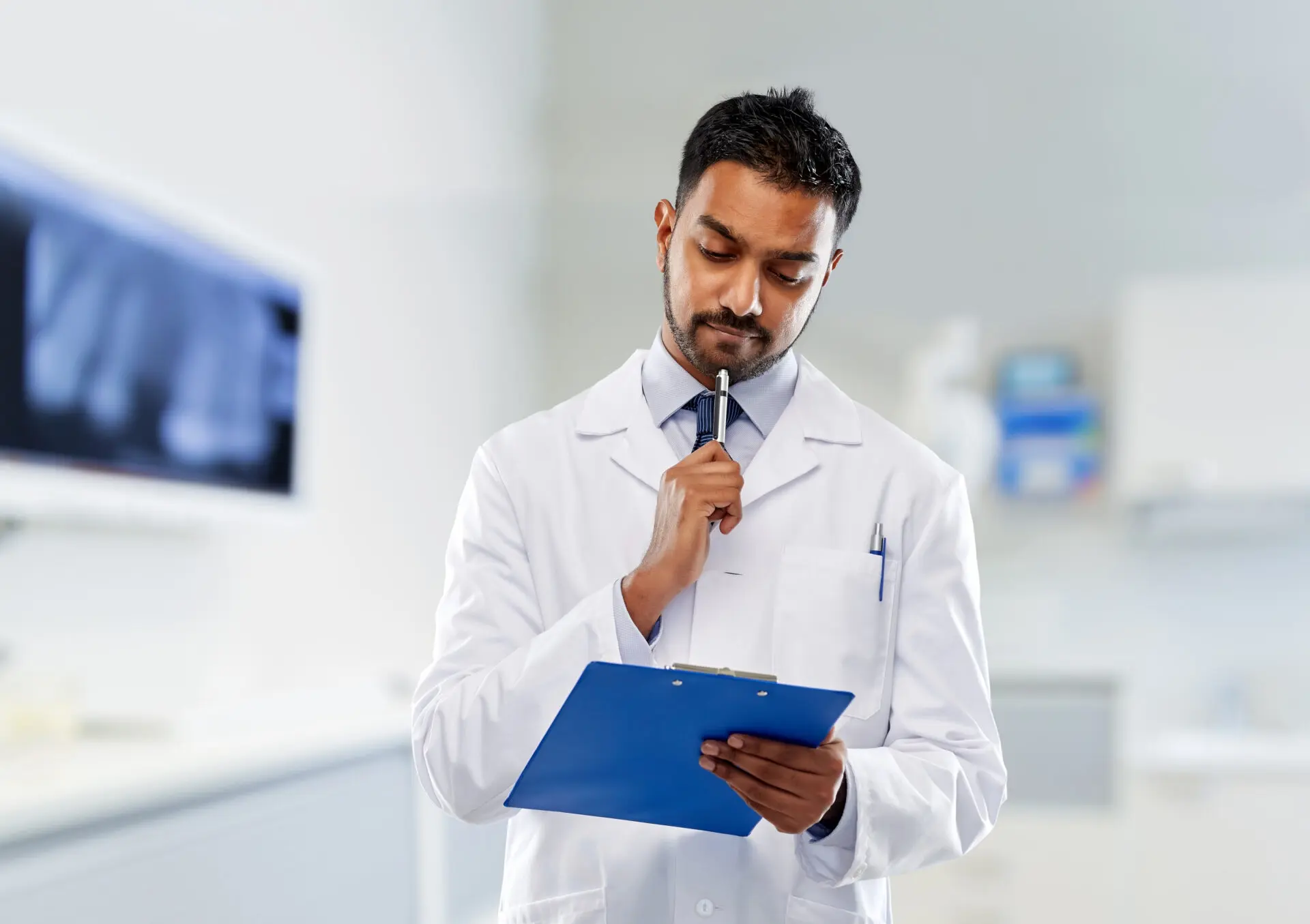 Indian male dentist in white coat holding a clipboard in a dental clinic, thinking about common dental myths and patient education.