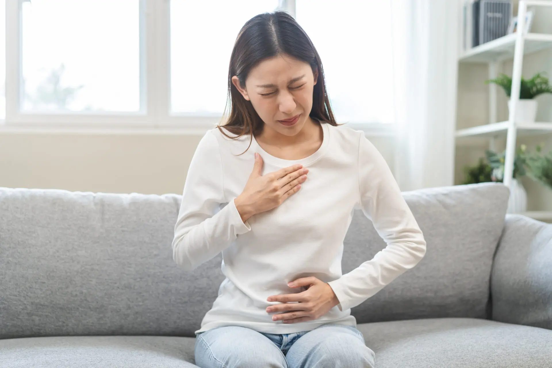 Asian woman holding her chest with stomach pain and heartburn, showing symptoms of acid reflux after eating, healthcare and medical concept.