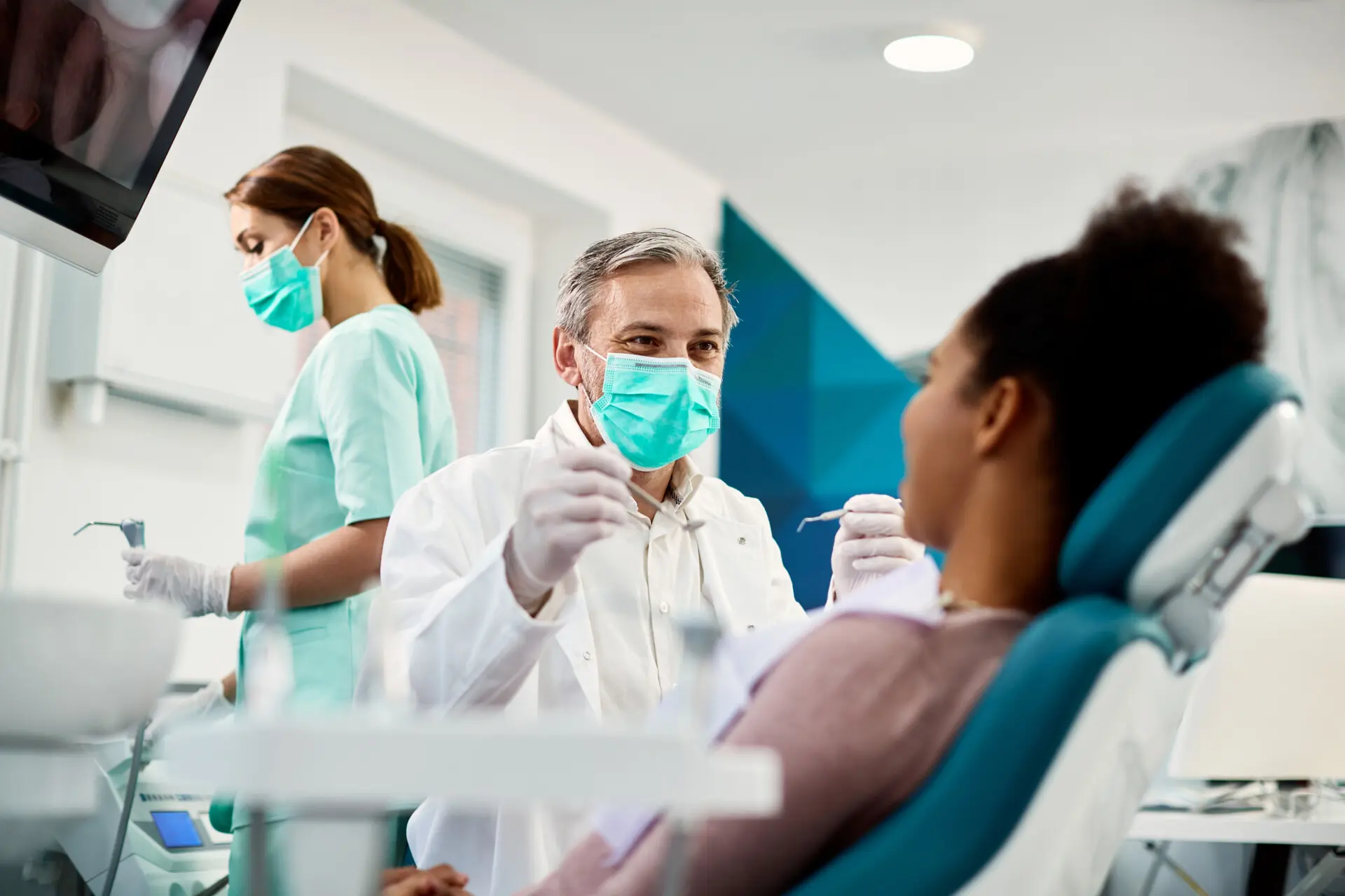 Smiling dentist with face mask talking to woman during emergency dental procedure at dental clinic.