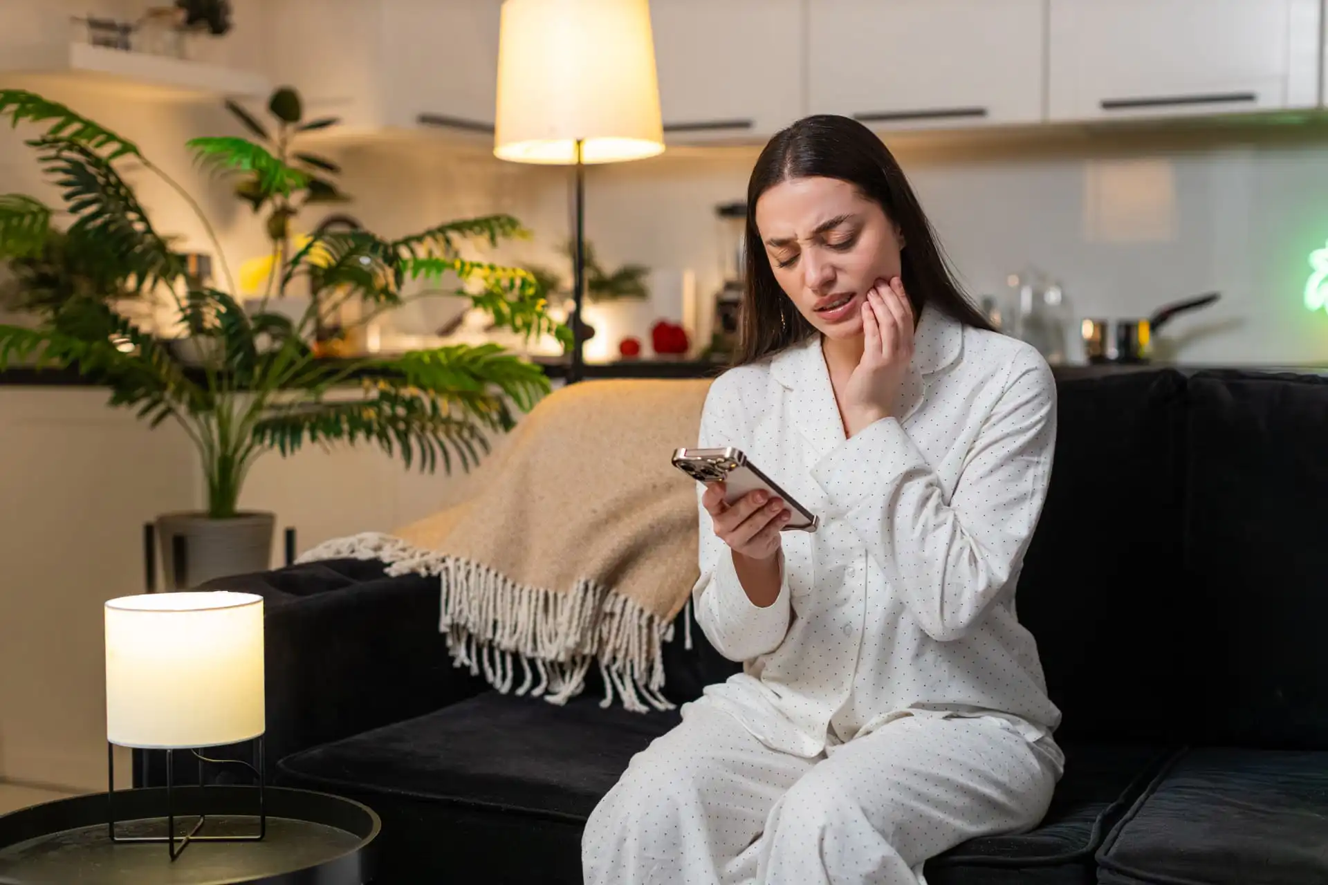 Woman sitting awake at night with tooth pain while looking at her phone, representing a nighttime toothache