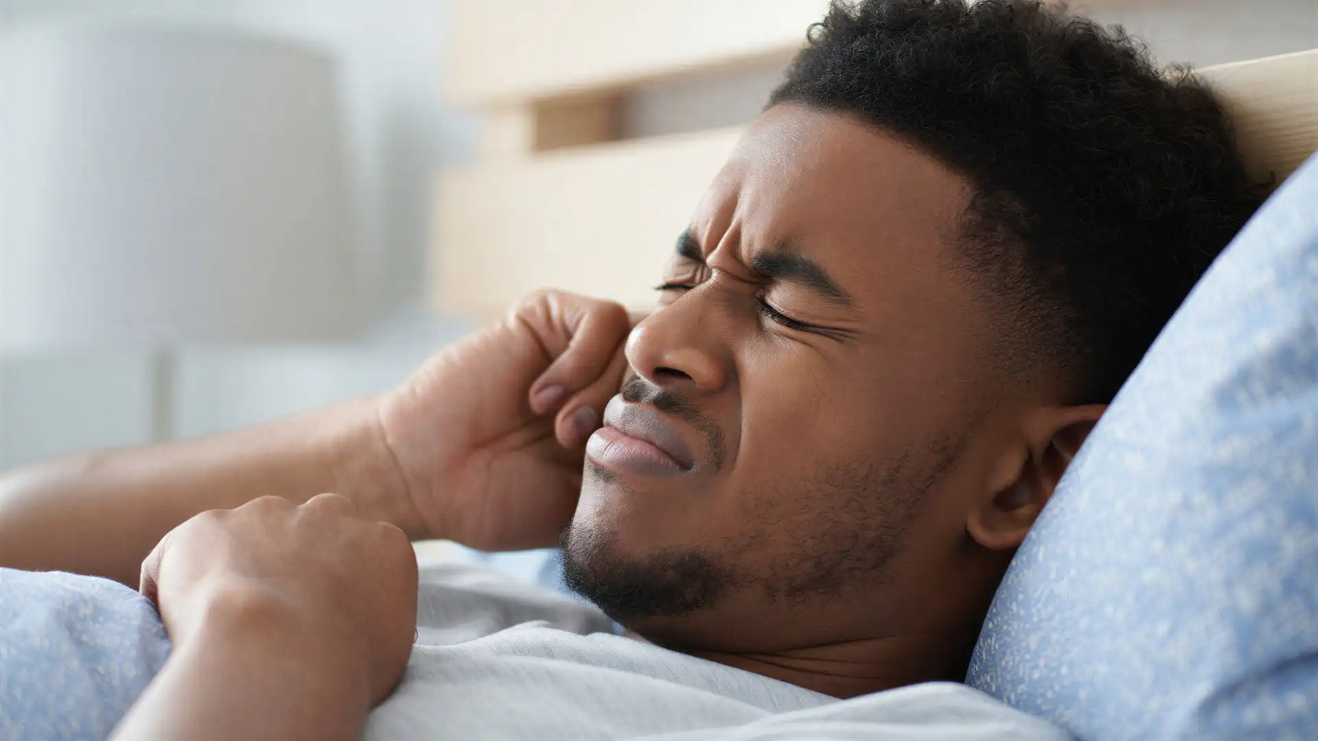 Man lying in bed while holding the side of his face near his ear in discomfort